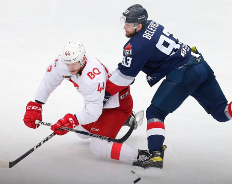 Игрок в красной форме с тростью борется за шайбу с игроком в синем на льду. Оба хоккеиста на полном ходу, напряженный момент матча. Видны детали экипировки и линии разметки