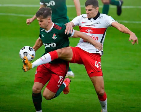 На изображении два футболиста в момент борьбы за мяч на поле. Один в форме зеленого цвета, другой — в белой с красными элементами. Оба активно движутся, пытаясь завладеть мячом