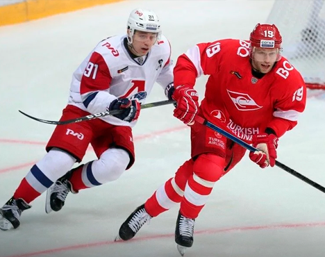 На изображении двое хоккеистов в игровой форме команд, один в красно-белой форме с номером 19, другой в бело-красно-синей с номером 91. Они соревнуются на льду, ведя борьбу за шайбу. Видны части хоккейной площадки