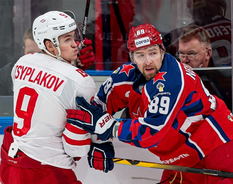 На изображении двое хоккеистов в игре: один в красно-синей форме, другой в бело-красной. Они борются у борта, рядом стоит судья. Атмосфера напряженная и динамичная