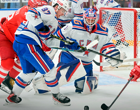 На изображении два хоккеиста в белых формах с синими и красными элементами защищают ворота. Один вратарь в полной экипировке ловит шайбу, другой игрок пытается выбить её из зоны. Форма команды — СКА