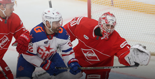 На изображении хоккейный матч: два игрока в красной форме противостоят игроку в синей. Вратарь в красной форме готовится поймать шайбу. Игроки сосредоточены на игре, лед чистый, сетка ворот видна на заднем плане